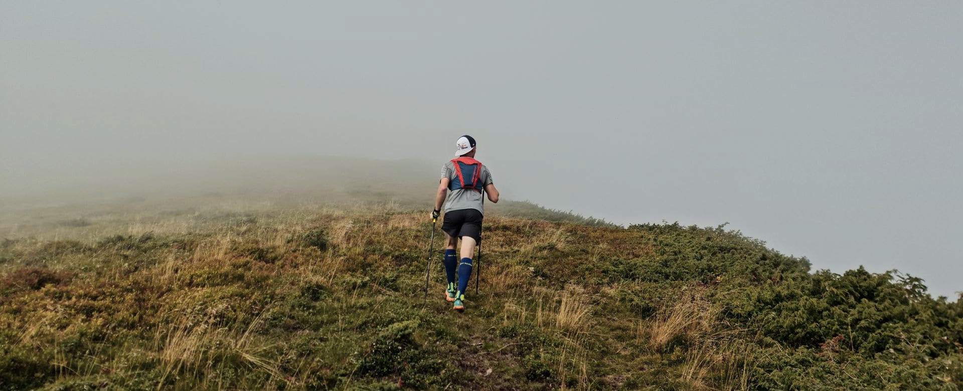 Trail ultrarunner with poles climbing a grassy ridge into the fog